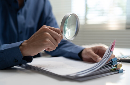 A professional male businessman uses a magnifying glass to carefully inspect a thick stack of paperwork. The auditor reviews financial documents and business contracts at his desk.