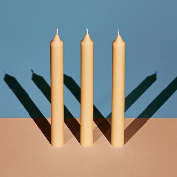 Three white taper candles standing upright with sharp parallel shadows on a two-tone background, for Christmas card layouts and festive banners