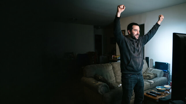 Excited man celebrating a victory while watching television in a dark living room at night