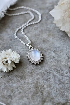 Elegant moonstone pendant necklace with delicate silver setting and clear stones, resting on textured gray stone with white flowers in the background