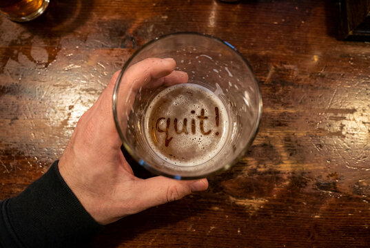 A hand holds an almost empty beer glass where leftover foam, wet residue, and tiny bubbles form the readable word quit! on the bottom.