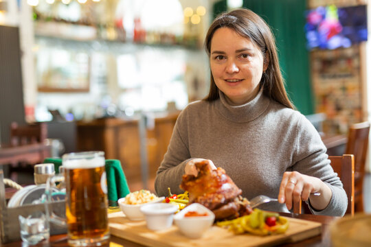 Smiling woman tourist enjoying traditional baked pork knuckle served with pickles, sauces and glass of beer in Viennese restaurant