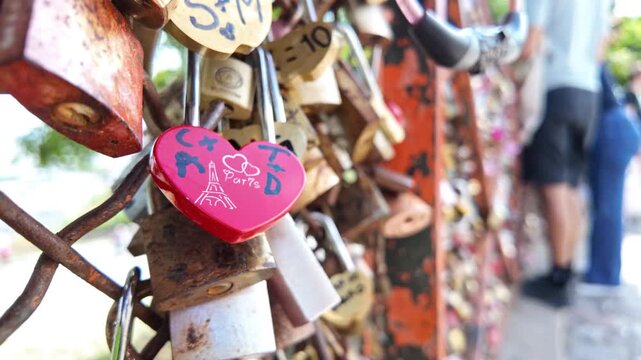 Red heart-shaped padlock marked with initials hangs among rusted love locks on a metal fence while tourists stand in the background.