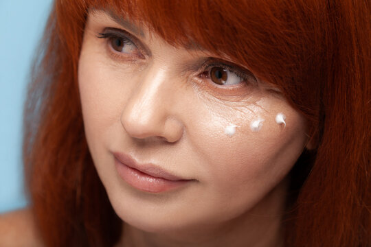 Middle aged woman applying three drops of moisturizing white face cream for daily skincare routine and anti-wrinkle treatment isolated on blue background