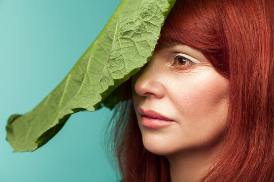 Middle aged woman hiding face behind large green leaf while performing organic skincare treatment and facial hydration procedure isolated on turquoise background