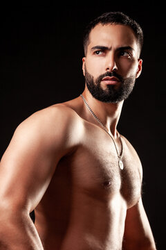 Shirtless fit man with sculpted pectoral and shoulder muscles in powerful three-quarter view with silver pendant necklace in dramatic lighting isolated over black background