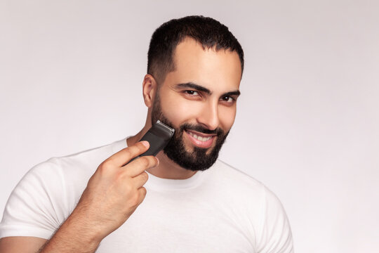 Smiling man grooming beard with professional trimmer holding modern electric clipper device wearing white cotton t-shirt posing isolated over gray background