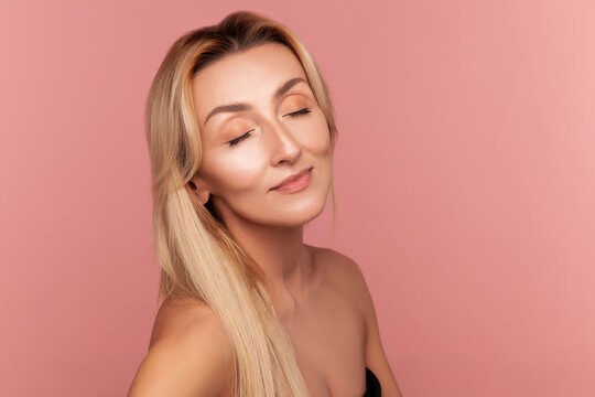 Serene blonde woman with closed eyes enjoying skin softness and face glow after professional aesthetic rejuvenation procedure isolated on pink background