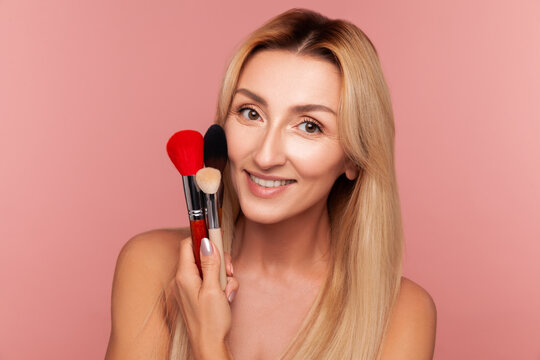 Smiling middle aged lady holding set of professional makeup brushes while preparing for beauty treatment isolated on pink background
