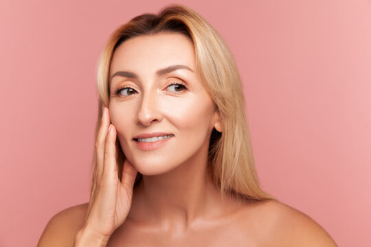 Radiant woman looking away and touching cheek with fingers for facial lifting and hydration concept isolated on pink background