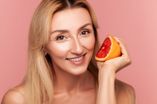 Radiant woman squeezing slice of red grapefruit near face, demonstrating refreshing citrus therapy and organic facial treatment isolated on pink background