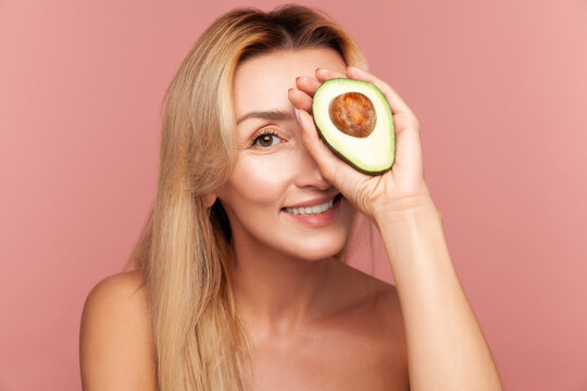 Playful woman covering eye with half of green avocado, showing organic ingredient for homemade face masks and nourishment isolated on pink background