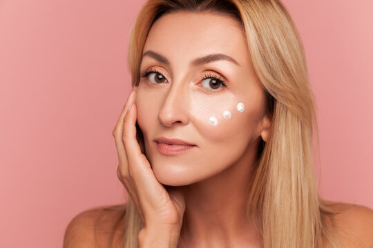 Mid-adult woman with blonde hair gently touching her face while posing with three moisturiser dots under eye isolated on pink background