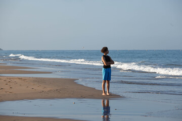 ​Young Man Standing Reflectively on Beach Looking Out at Ocean, Low Angle Shot. A barefoot young man with curly dark hair stands alone on a sandy beach. © NataSao