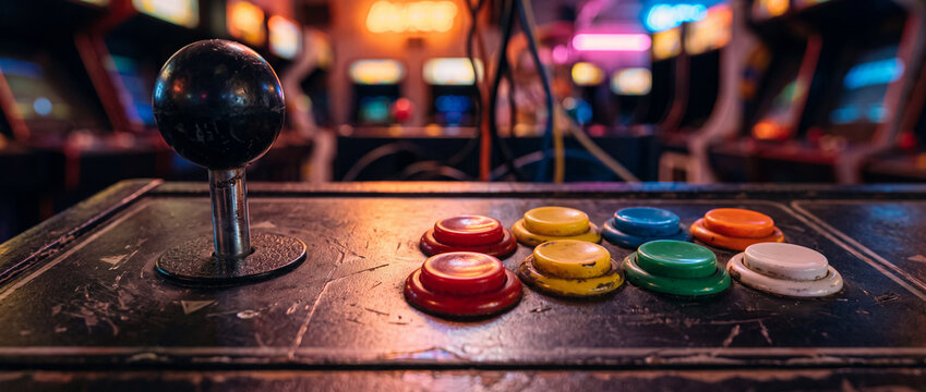 Worn arcade joystick and multicolored action buttons on a vintage game console, framed by neon-lit arcade machines and blurred lights to evoke nostalgia and retro gaming energy.