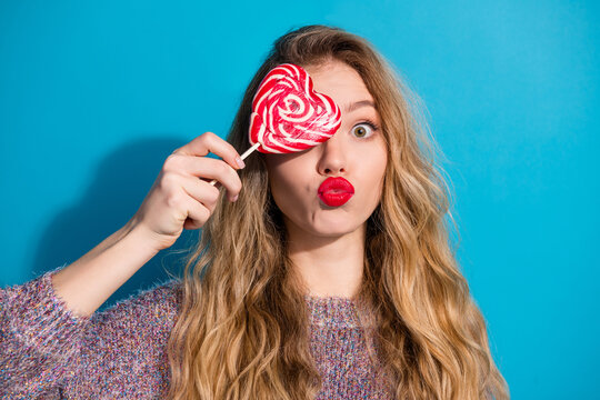 Young woman with heart lollipop covers eye puckered lips blue studio background fashion playful lifestyle