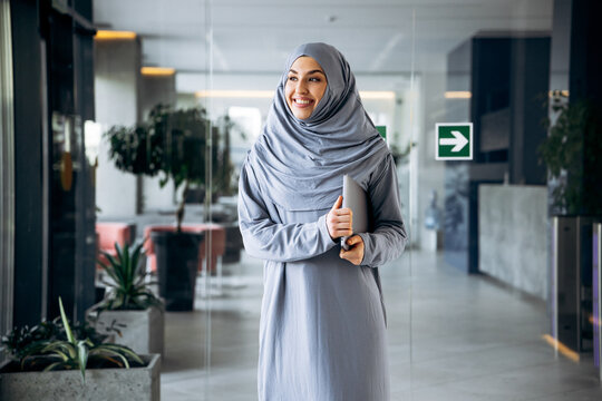 Muslim woman in hijab carrying a modern laptop, smiling confidently and looking away from camera in a bright office environment
