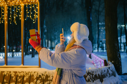 Woman standing in a snowy winter park at night. Holding a wrapped christmas gift. Using her phone to take a selfie under the warm glow of decorative string lights. Capturing and sharing holiday cheer
