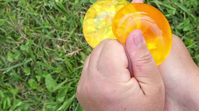 Two hands clutch yellow and orange translucent gel-like slimes on green grass, gradually squeezing and changing their shape as the balloons bend, bulge and coalesce outdoors in daylight.