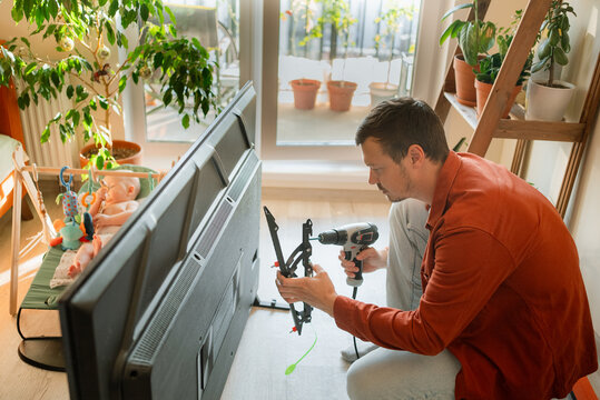 Man installing a tv on wall brackets in a bright apartment, holding a drill while a baby watches from a bouncer, showcasing home improvement and family life