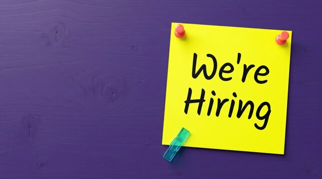 Bright yellow sticky note with the text we are hiring pinned to a dark purple wooden texture surface background board