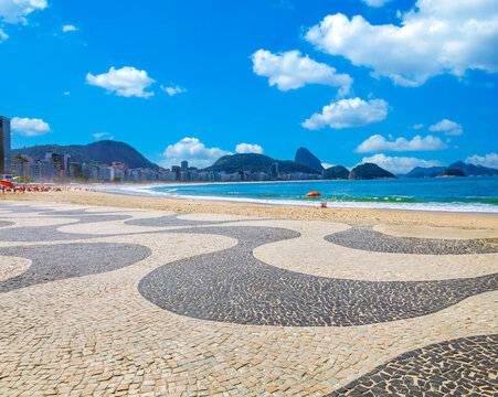 Iconic Copacabana beach promenade in Rio de Janeiro, Brazil. Sunny day featuring the famous wave mosaic sidewalk, blue ocean, Sugarloaf mountain, and clear sky.