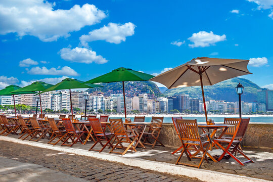 Outdoor cafe seating at Copacabana Fort, overlooking the beach, city skyline, and mountains on a sunny day in Rio de Janeiro, Brazil.