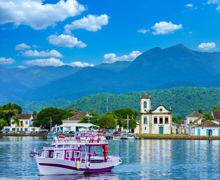 Scenic coastal view of historic Paraty, Brazil, with a boat, church, and lush mountains under a bright blue sky.