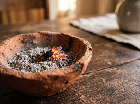 Burning incense stick in a rustic clay bowl with ash on wooden table