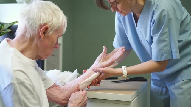 Gentle nurse unwraps medical bandage from senior patient hand in clinic. Caring staff helps convalescent man recover after fracture with tender care.