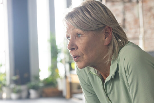 Senior woman leaning forward wearing light green zip-up top in bright room with plants, copy space