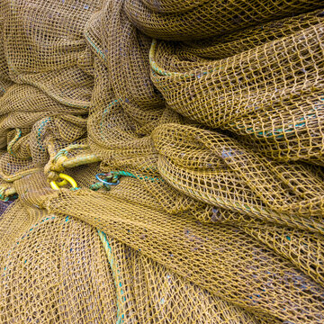 A detailed close-up of piled, textured yellowish-brown commercial fishing nets on a deck, featuring complex mesh patterns, knots, and a visible yellow hardware ring.