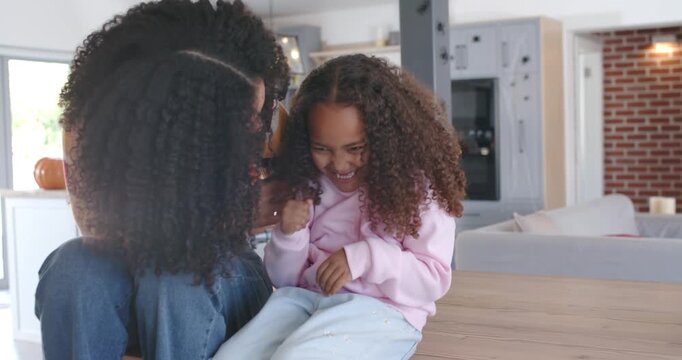 African American mother leaning in, tickling daughter in pink hoodie on kitchen island, laughing