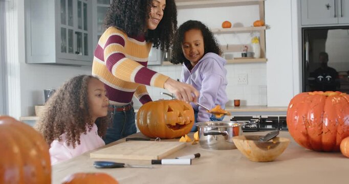 African American mom with two child girls seeing jack-o'-lantern cleaning pulp into bowl on island