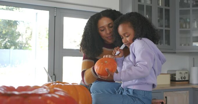 African American mother handing pumpkin, guiding child tracing with pen on kitchen counter for fall
