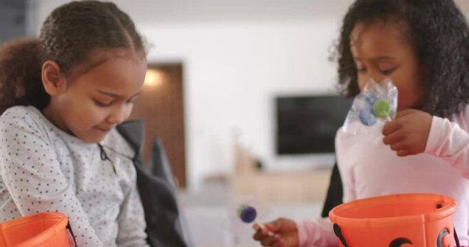 African American girls trading lollipops, in black capes, filling orange buckets with candy at home