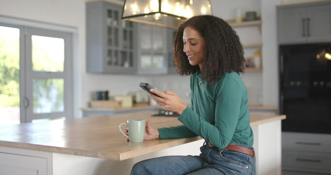 African American woman sitting on stool at island, reading phone then speaking into it holding mug