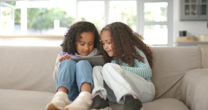 Two African American child girls sitting on sofa, settling with tablet, tapping screen and learning