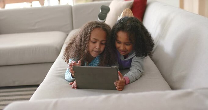 African American girls siblings lying on gray sofa sharing tablet case, tapping as content changing