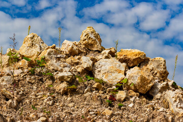 Heap of yellow limestone rocks in an open-pit quarry with sparse wild plants growing against a vibrant blue sky with scattered soft white clouds in sunny daylight conditions © PhotoChur