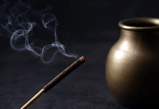Burning incense stick with rising smoke next to a golden ceramic pot on dark background