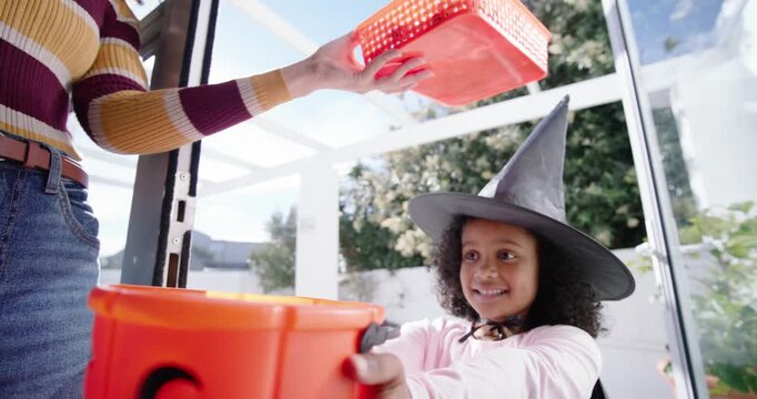 African American girl in witch hat holding pumpkin pail at door, adult pouring candy for Halloween