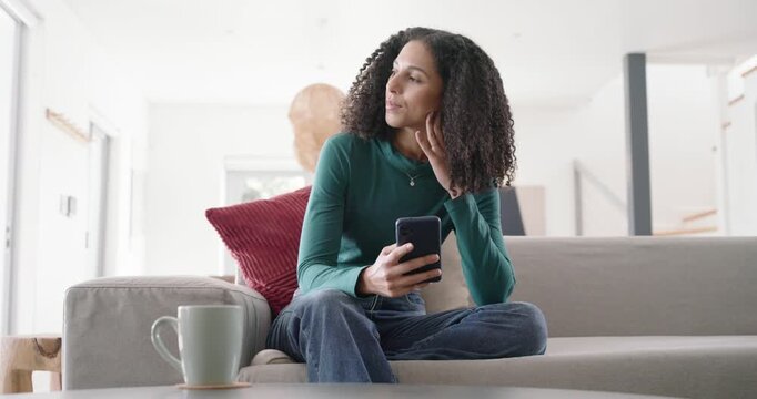 African American woman checking puzzling phone message, scrolling, smiling, sipping mug on sofa