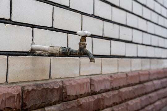 Rusty outdoor faucet on tiled wall shows weathered plumbing in close up. Old metal tap has white handle, corroded pipe, and brick masonry backdrop with stains, grime, fittings, and surface texture.
