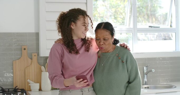 Youth woman sharing smartphone, senior woman leaning in, smiling and laughing at kitchen counter