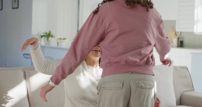 African American women, one in pink sweatshirt approaching, hugging other on sofa at home