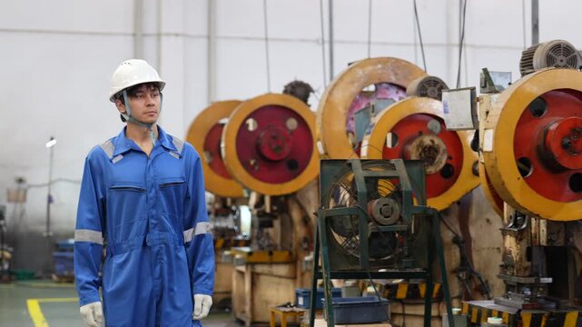 Industrial worker wearing protective gear, helmet and blue coverall inspecting mechanical press equipment beside heavy machinery, with technician and engineer supporting safe manufacturing workshop