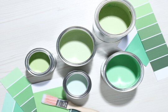 Metal cans with paint in shades of green, brush and color palettes on white wooden table, flat lay