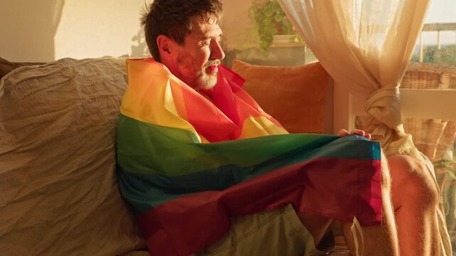 mature queer man wearing lipstick and holding a gay pride flag while smiling during sunset