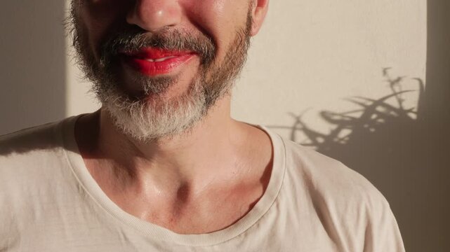 Close-up of a smiling bearded non-binary person wearing bright red lipstick in the sunlight